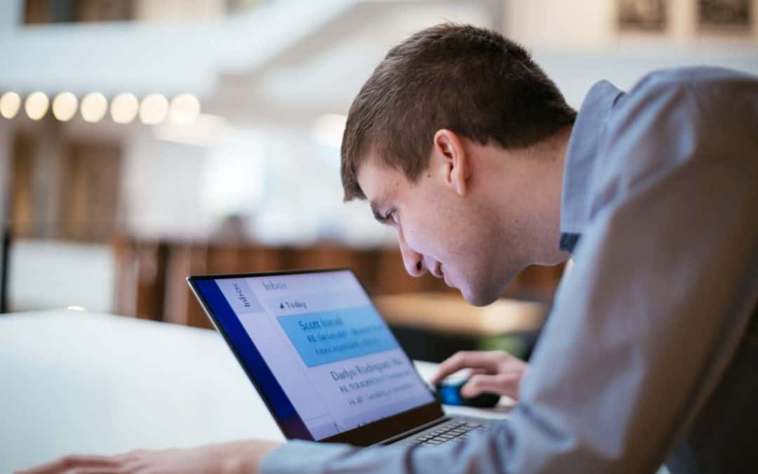 Eine männlichePersonsitzt am Schreibtisch mit BLick auf den Bildschirm eines Notebooks